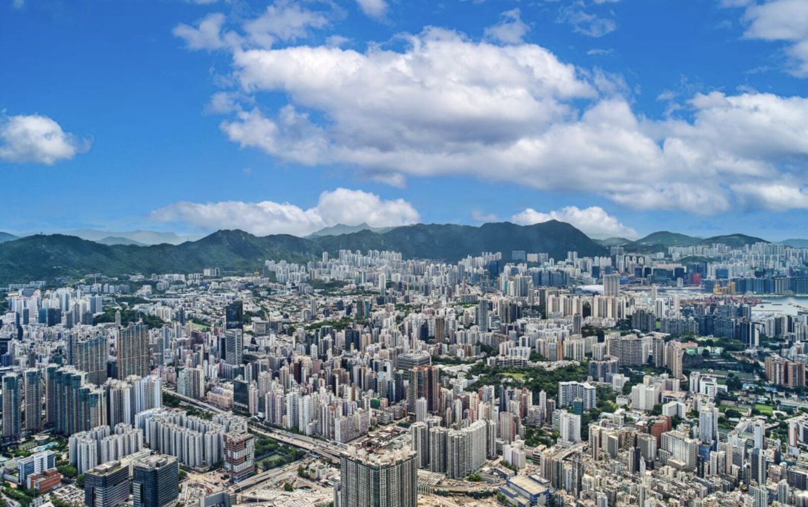 An aerial view over the Kowloon skyline and hills beyond, as seen from high above the city