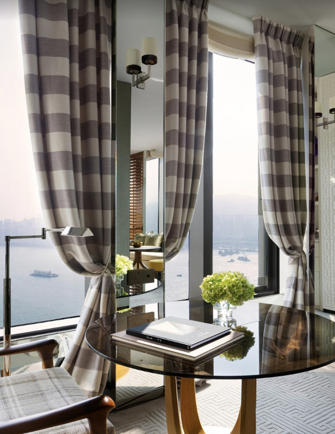 A round table by the window in a room at Rosewood Hong Kong, with striped curtains and a harbour view