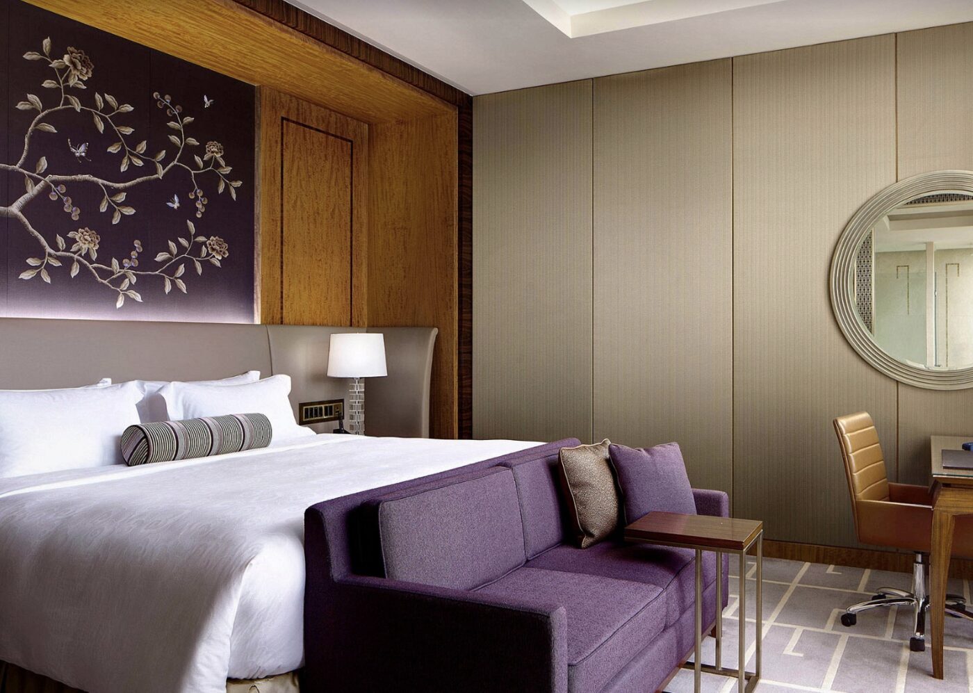 A bedroom at The Royal Garden with a plum sofa and a round mirror over the desk